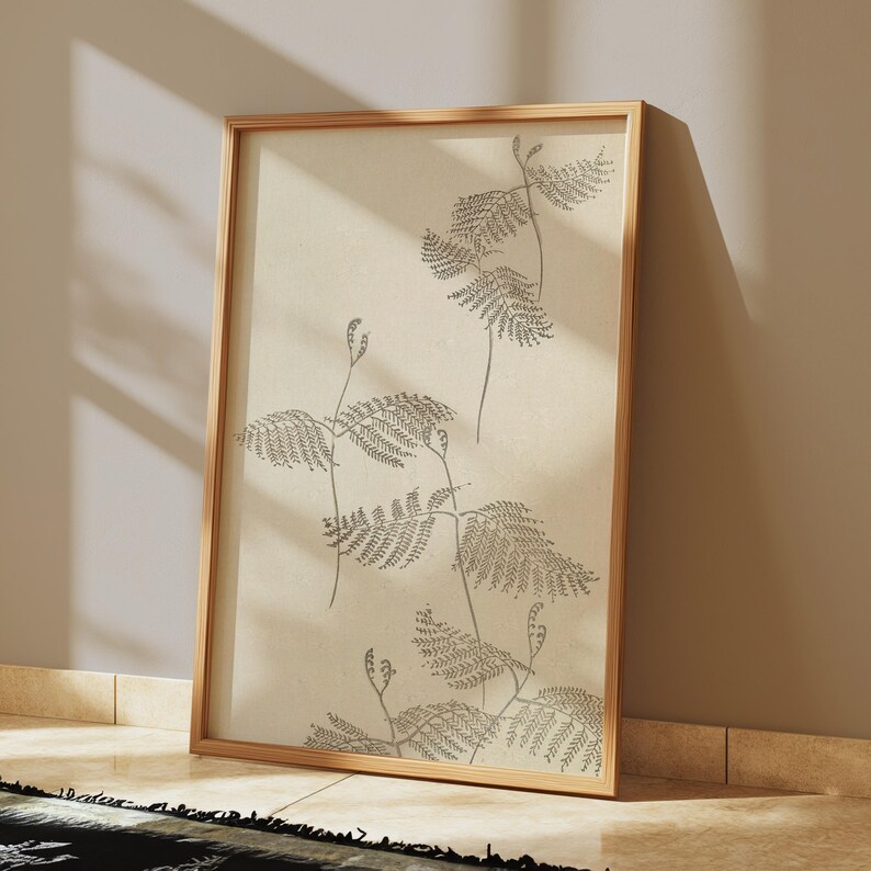 May include: A framed botanical print featuring a detailed illustration of fern-like plants in shades of gray. The artwork is set against a cream-colored background and encased in a natural wood frame. The print is leaning against a neutral-toned wall.