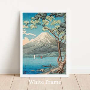 Op de afbeelding: Een ingelijste prent van een Japans landschap met de Fuji. Het kunstwerk toont een serene meer met boten, een boom met witte bloesems en een besneeuwde berg onder een blauwe hemel. De lijst is wit, en de tekst "White Frame" staat onderaan.
