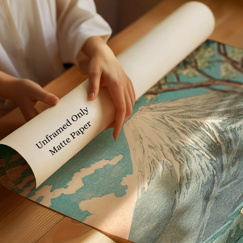 Op de afbeelding: Een opgerolde, ongerijmde matte papieren print wordt door handen vastgehouden. De print toont een landschap met een berg en bomen, met de tekst "Unframed Only Matte Paper". De print heeft blauwe, witte en groene kleuren.