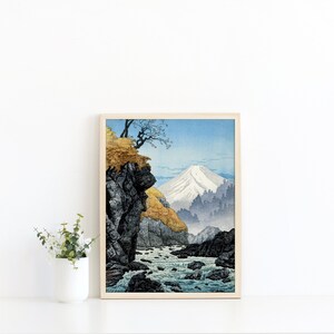 Pu&ograve; includere: Una stampa incorniciata di un paesaggio giapponese con una montagna con una cima innevata, un fiume impetuoso e una scogliera rocciosa con un piccolo albero che cresce in cima. La stampa &egrave; in una cornice di legno chiaro.