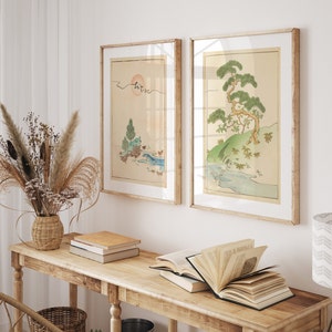 May include: Two framed prints of Japanese-style landscape paintings with a light wood frame. The prints are hanging on a white wall above a wooden console table with books and a vase of dried flowers.
