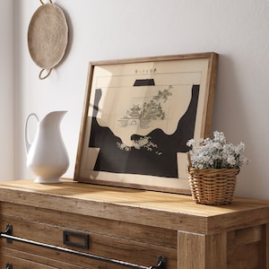 May include: A framed black and white print of a Japanese map with the text "Suita-ku" in the top right corner. The print is hanging on a white wall above a wooden dresser with a white pitcher and a wicker basket of white flowers.