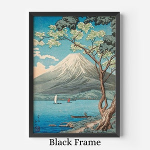 Op de afbeelding: Ingelijste kunstdruk met een Japans landschap met de berg Fuji, een meer en boten. Het kunstwerk zit in een zwarte lijst. De lucht is blauw met witte wolken en de berg is besneeuwd. Een boom met groene bladeren en witte bloemen staat op de voorgrond.