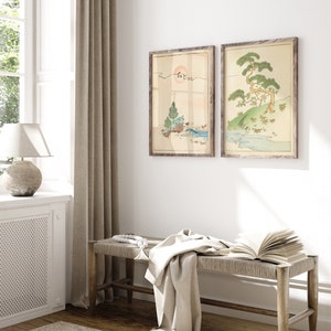 May include: Two framed prints of Japanese-style artwork depicting a landscape with trees, water, and a sun. The prints are hung on a white wall above a wooden bench with a white throw blanket and a book.