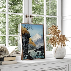 Pu&ograve; includere: Una stampa incorniciata di un paesaggio giapponese con una montagna, un fiume e alberi. La stampa &egrave; in una cornice di legno ed &egrave; appesa a un muro bianco.