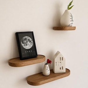 May include: Three wooden floating shelves with decorative items. A framed black and white moon print, a small white vase with a red flower, a white house-shaped candle holder, and a white vase with a green plant. The shelves are oval-shaped and made of wood.