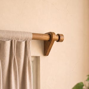 May include: Beige linen curtains suspended from a wooden curtain rod with decorative brackets. The rod and brackets are light brown, and the curtains have vertical pleats. The background is a light beige wall.