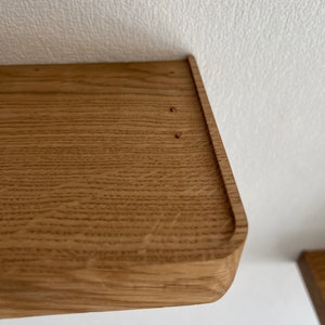 May include: Close-up of a wooden shelf with a rounded edge. The shelf is made of light brown wood with a natural grain pattern. There are two small holes in the top of the shelf.