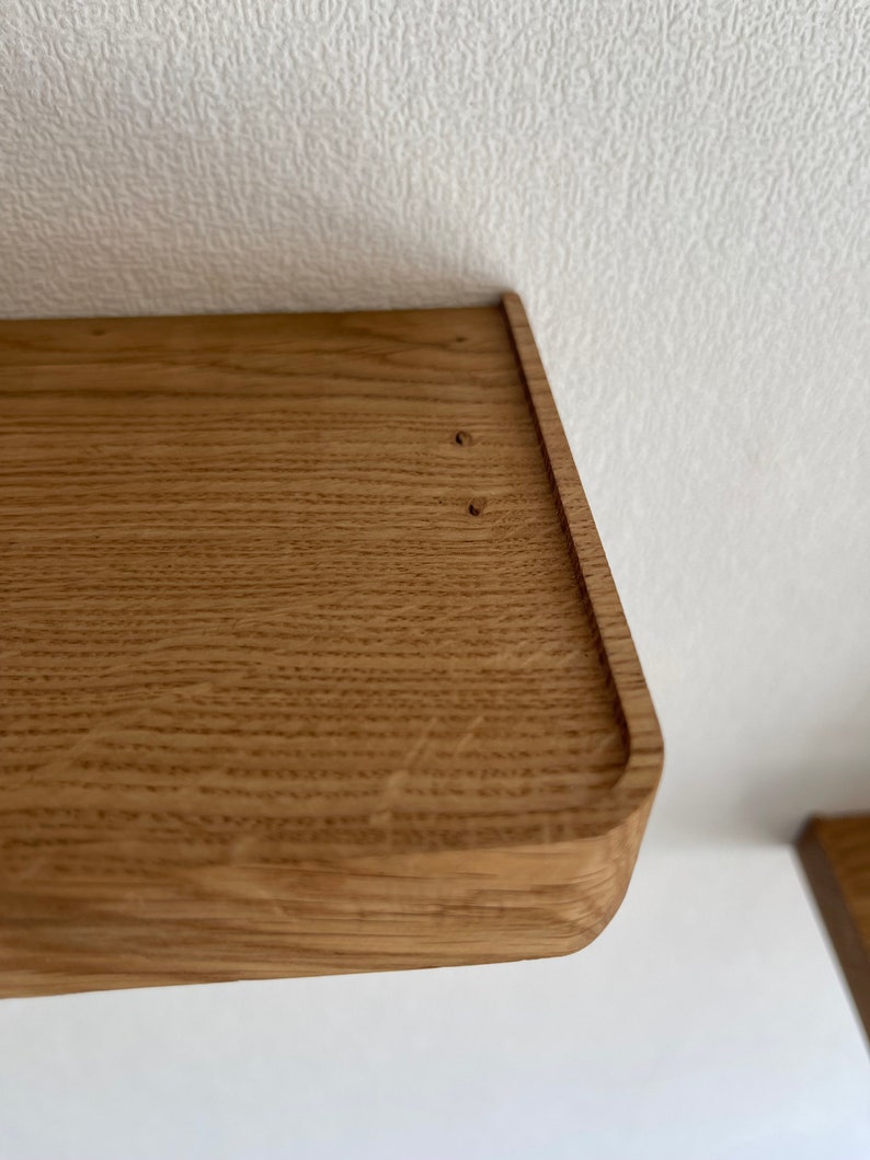 May include: Close-up of a wooden shelf with a rounded edge. The shelf is made of light brown wood with a natural grain pattern. There are two small holes in the top of the shelf.