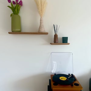 May include: Two wooden shelves with a green vase with purple tulips, a white vase with dried pampas grass, a brown bottle with black sticks, and a black rectangular object on the top shelf. A record player with a black turntable and a clear lid is on a black stand below the shelves.