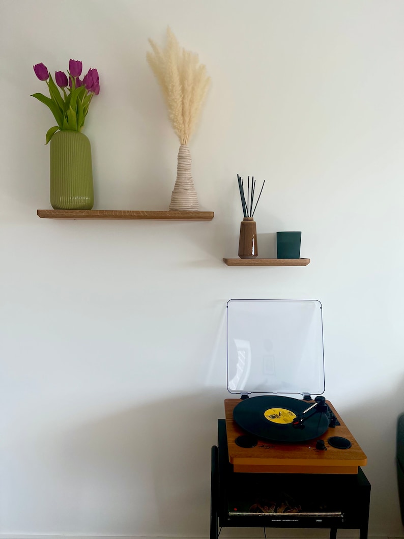 May include: Two wooden shelves with a green vase with purple tulips, a white vase with dried pampas grass, a brown bottle with black sticks, and a black rectangular object on the top shelf. A record player with a black turntable and a clear lid is on a black stand below the shelves.
