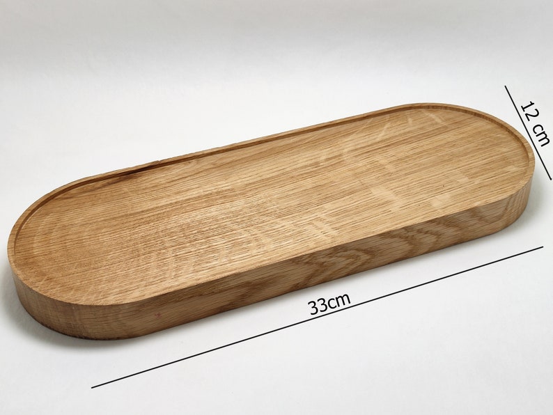 May include: A light brown, oval-shaped wooden tray. The tray is 13 inches long and 4.7 inches wide. The tray has a raised edge and is made of wood. The tray is empty and sits on a white surface.