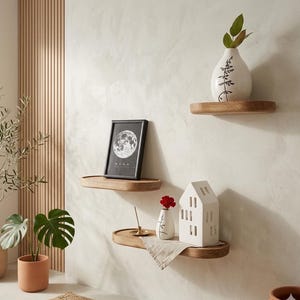 May include: Three wooden floating shelves mounted on a light-colored wall. The shelves display decorative items, including a framed picture with a moon design, a white ceramic house, and small vases with plants. The shelves are made of light-colored wood.