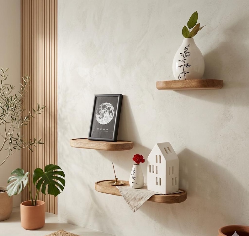 May include: Three wooden floating shelves mounted on a light-colored wall. The shelves display decorative items, including a framed picture with a moon design, a white ceramic house, and small vases with plants. The shelves are made of light-colored wood.