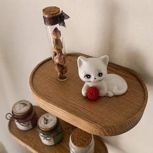May include: A white ceramic cat figurine with black eyelashes and a red ball, resting on a wooden shelf. A glass vial filled with dried roses and small jars with cork lids are also on the shelf. Decorative home decor.