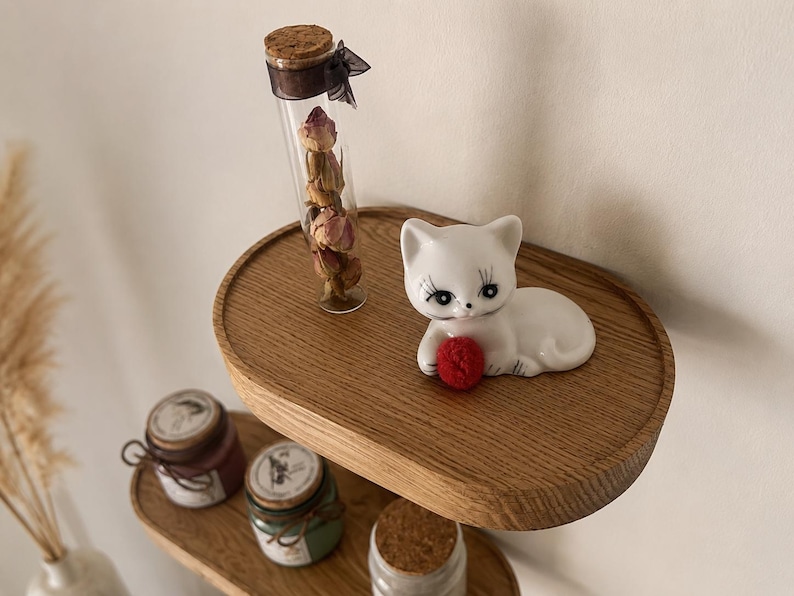 May include: A white ceramic cat figurine with black eyelashes and a red ball, resting on a wooden shelf. A glass vial filled with dried roses and small jars with cork lids are also on the shelf. Decorative home decor.