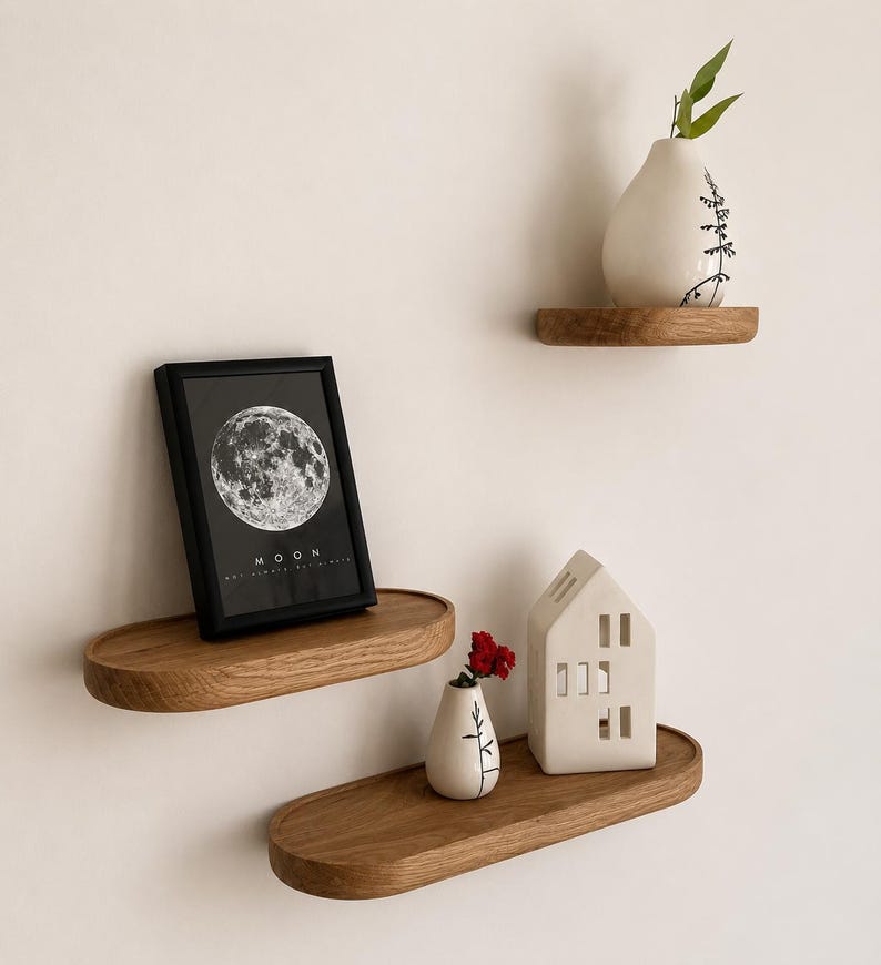 May include: Three wooden floating shelves with decorative items. A framed black and white moon print, a small white vase with a red flower, a white house-shaped candle holder, and a white vase with a green plant. The shelves are oval-shaped and made of wood.