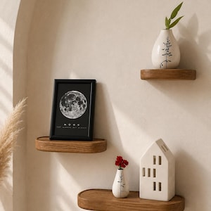 May include: Three wooden floating shelves with decorative items. A framed black and white moon print, a white vase with a tree design, and a small white house-shaped candle holder. The shelves are mounted on a neutral-colored wall.