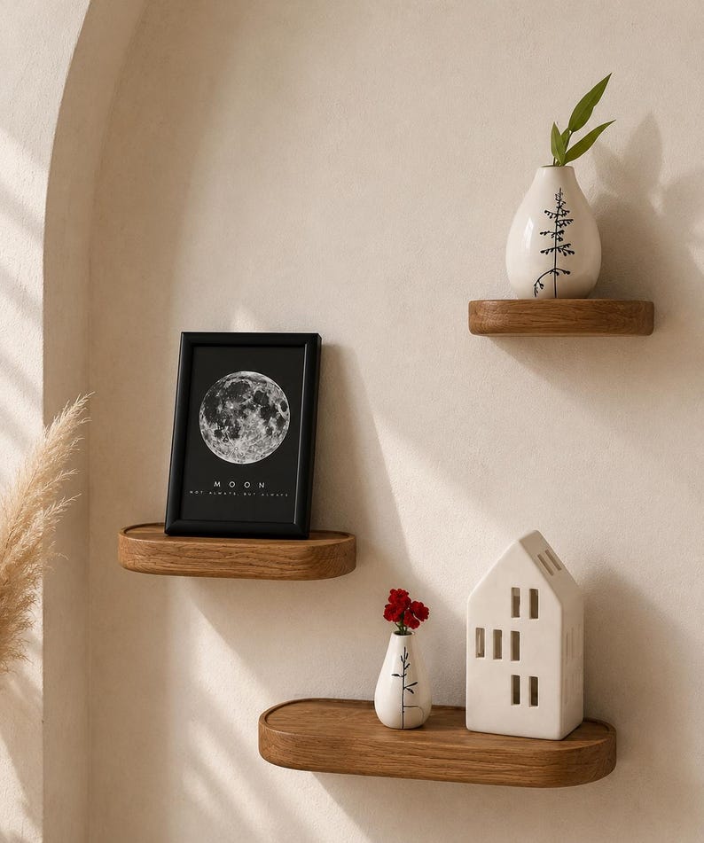 May include: Three wooden floating shelves with decorative items. A framed black and white moon print, a white vase with a tree design, and a small white house-shaped candle holder. The shelves are mounted on a neutral-colored wall.