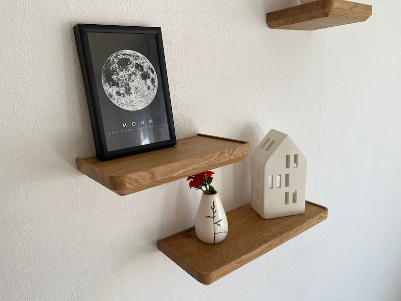 May include: Two wooden shelves with rounded edges, mounted on a white wall. The top shelf has a black framed print with a white moon graphic and the text "MOON". The bottom shelf has a white ceramic house-shaped candle holder and a white vase with a red flower.
