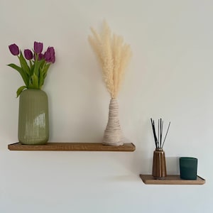 May include: Two wooden shelves with three decorative items. The top shelf has a green vase with purple tulips and a white ceramic vase with dried pampas grass. The bottom shelf has a dark green glass diffuser and a black diffuser.