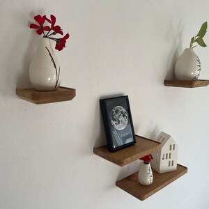 May include: Three wooden shelves mounted on a white wall. The top shelf has a white vase with red flowers. The middle shelf has a black framed picture with a moon image and the word "Moon" printed below. The bottom shelf has a white ceramic house and a white vase with red flowers.