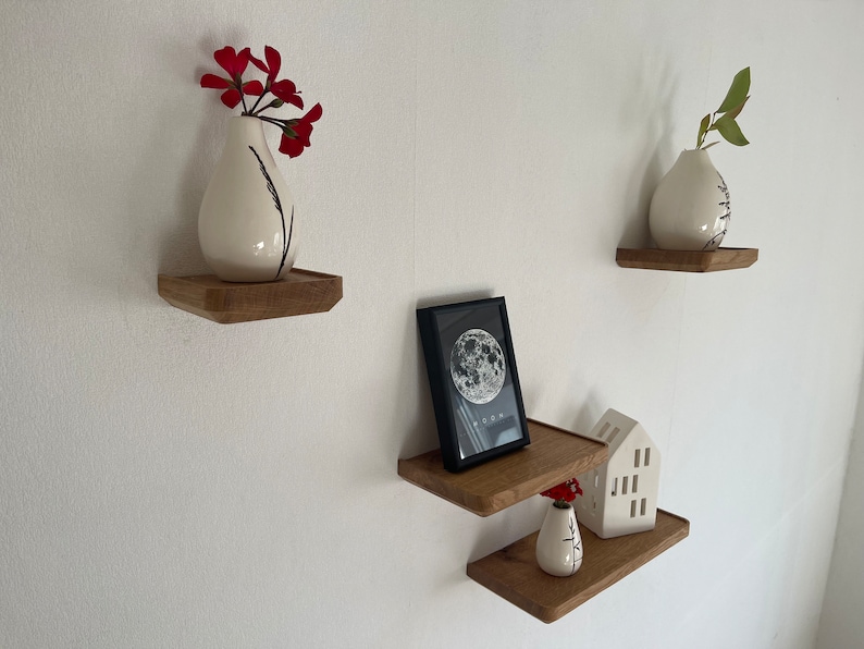 May include: Three wooden shelves mounted on a white wall. The top shelf has a white vase with red flowers. The middle shelf has a black framed picture with a moon image and the word "Moon" printed below. The bottom shelf has a white ceramic house and a white vase with red flowers.