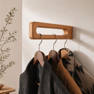 Op de afbeelding: Een houten wandkapstok met drie metalen haken. De kapstok is gemaakt van lichtgekleurd hout en heeft een rechthoekige vorm met een uitgesneden handvat. Drie houten kleerhangers houden jassen van verschillende kleuren en patronen vast.