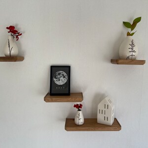 May include: Three wooden shelves with white ceramic vases, a black framed print of the moon, and a white ceramic house figurine. The vases are decorated with black floral designs. The shelves are mounted on a white wall.
