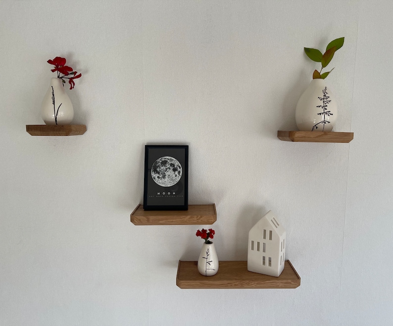 May include: Three wooden shelves with white ceramic vases, a black framed print of the moon, and a white ceramic house figurine. The vases are decorated with black floral designs. The shelves are mounted on a white wall.