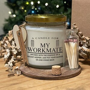 May include: A white candle in a glass jar with a gold lid. The candle has a black label that reads "A candle for my workmate. Infused with the essence of a wicket keeper. Just put your gloves on and stand back." The candle is on a wooden base with a small glass bottle of matches.