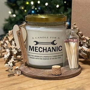 May include: A candle for a mechanic in a glass jar with a gold lid, featuring the text "A CANDLE FOR A MECHANIC". The candle sits on a wooden coaster with a small glass jar of matches and a cork.