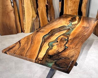 Op de afbeelding: Een grote houten tafel met een live edge en een turquoise epoxyhars inlay. De tafel heeft een zwart metalen X-vormig onderstel.