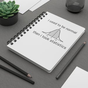 May include: A white spiral-bound notebook featuring a black graphic of a normal distribution curve and the text "i used to be normal then i took statistics" printed on the front cover. The notebook has a black metal coil binding along the side.