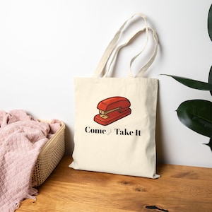 May include: Cream-colored tote bag featuring a red stapler graphic and the text "Come and Take It" in black. The bag has two handles and is made of a light-colored fabric. The stapler is a classic office supply.