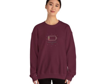 Puede incluir: Una sudadera burdeos con un gráfico de batería baja y el texto "SOCIAL BATTERY 1%" en un diseño circular. La sudadera es de color liso y tiene cuello redondo.