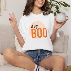 May include: A white short-sleeved t-shirt featuring a graphic design with a light peach-coloured circle background. The text "hey" is written in black cursive script above the word "BOO" in large, bold, orange sans-serif capital letters.