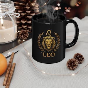 May include: Black ceramic mug featuring a gold Leo zodiac design. The design includes a lion's head, the Leo symbol, and the word "LEO" in gold. The mug has a black handle and is filled with a dark liquid.