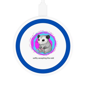 Pode incluir: Um carregador sem fio branco com um design de anel azul. O centro apresenta uma ilustração de desenho animado de um gambá dentro de um redemoinho rosa e azul. O texto "softly accepting the void" está abaixo.