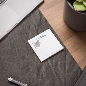 May include: A white square notepad with a black spider illustration and the name "Ashley" in script. The notepad is on a dark gray surface, with a laptop and a pen visible in the background.