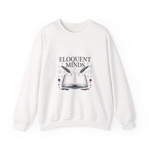 May include: White sweatshirt with the text "ELOQUENT MINDS" above an open book illustration with two quills. The book has red, green, and black accents. The design is surrounded by small symbols.
