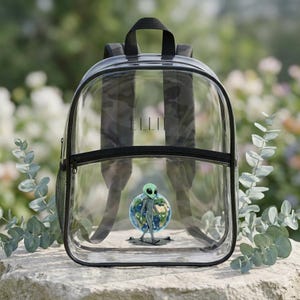 May include: Clear backpack with black trim, straps, and handle. The backpack features a zipper closure and a side mesh pocket. A graphic of a green alien standing on a globe is visible on the front. The name "ELLIE" is printed on the front.