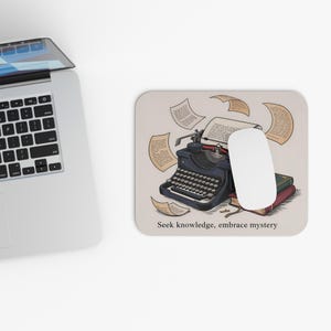 May include: A beige mousepad featuring a vintage typewriter illustration with flying papers and a white mouse. The mousepad has the text "Seek knowledge, embrace mystery" at the bottom. A laptop is visible in the background.