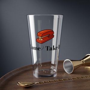 May include: A clear pint glass with a red and orange stapler graphic and the words "Come and Take" printed in black. A gold bar spoon and jigger are on a wooden tray.