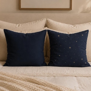 Puede incluir: Dos cojines cuadrados sobre una cama. Uno es de color azul marino liso y el otro es azul marino con un patrón de estrellas doradas. Están colocados frente a dos almohadas más grandes de color crema sobre una cama con un edredón crema.