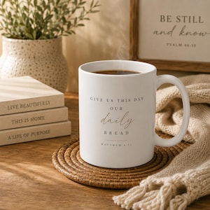 May include: A white ceramic mug with a handle, featuring the black and brown printed text GIVE US THIS DAY our daily BREAD and MATTHEW 6:11. The mug contains a dark liquid with visible steam rising from the top, resting on a round woven coaster.