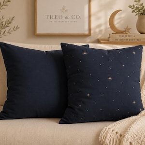 May include: Two square throw pillows in a deep navy blue color. One pillow is solid navy blue, while the other features a repeating pattern of small gold stars and larger four-pointed starbursts scattered across the dark navy blue fabric surface.
