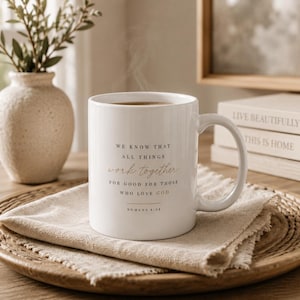 May include: A white ceramic mug with a handle, featuring the black and gold printed text: We know that all things work together for good for those who love God, Romans 8:28. The mug contains a dark liquid and is placed on a folded beige linen napkin.