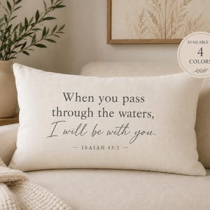 May include: A rectangular off-white throw pillow featuring the black printed text: When you pass through the waters, I will be with you. — ISAIAH 43:2 —. A circular badge in the upper right corner states: AVAILABLE IN 4 COLORS.
