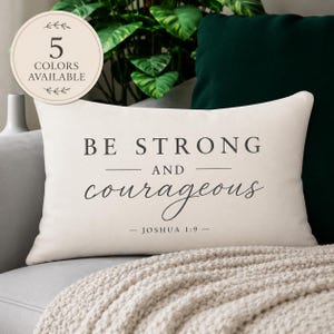 May include: A rectangular cream-colored throw pillow featuring dark gray text that reads BE STRONG AND courageous JOSHUA 1:9. A circular badge in the upper left corner displays the text 5 COLORS AVAILABLE with decorative leaf accents.